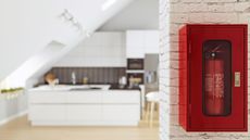 A small red fire extinguisher in a red, glass front wall-mounted box. A home kitchen blurry in the background