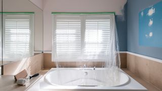 bathroom mid renovation with plastic over shutters and sunken bath, partly painted walls, paint roller and sandpaper resting on vanity unit
