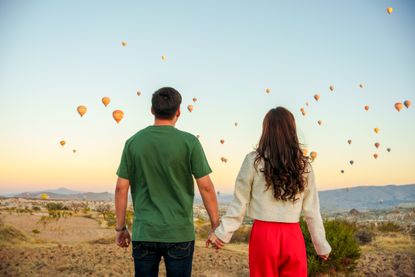 A couple holding hands while watching hot air balloons flying in the sky.