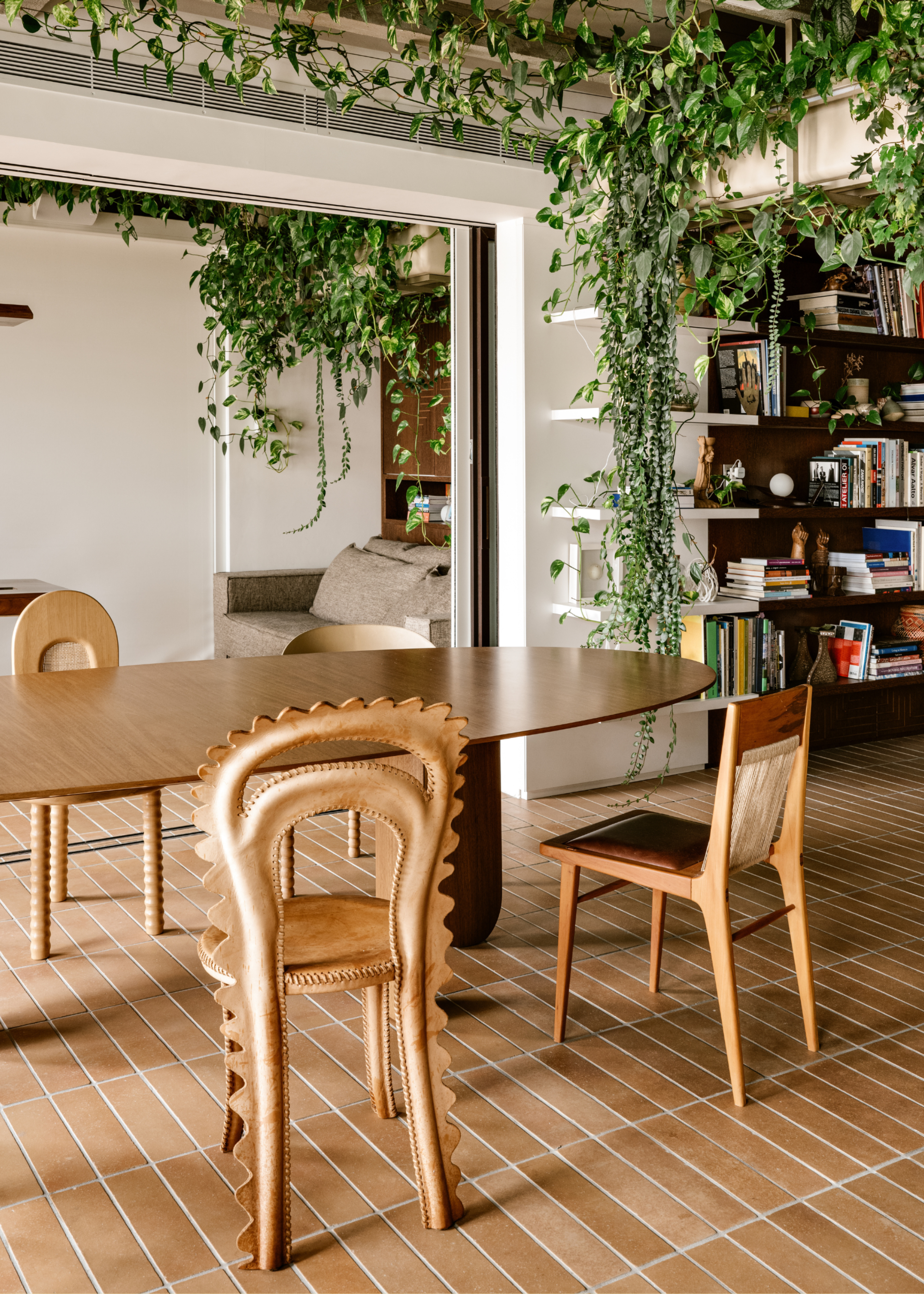 A dining space with a large wood table, mismatched chairs, green trailing plants, and a stacked book shelf