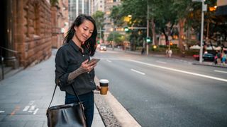 Bytte mobilabonnement: En ung kvinne med kaffekopp i hånden ser på mobilen sin på et fortau i en storby