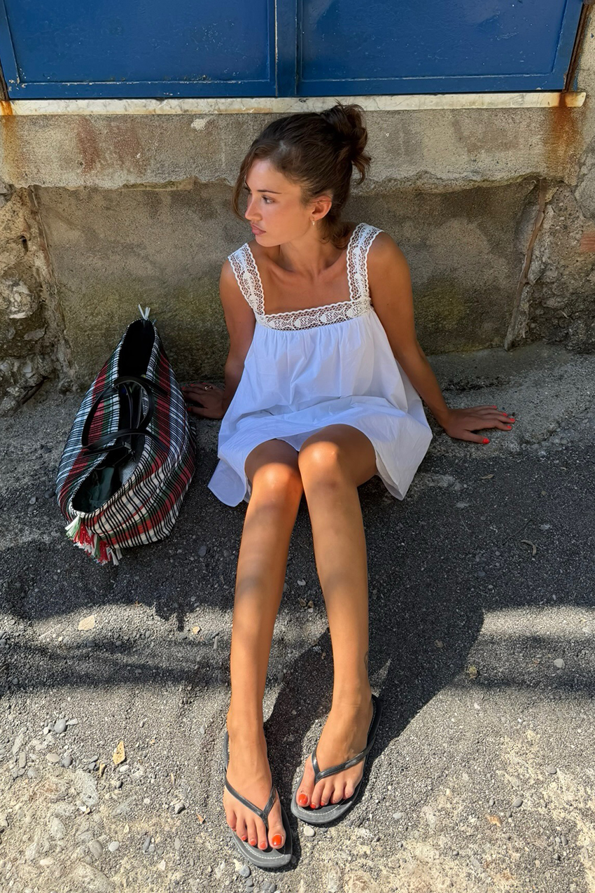 a woman wearing a white babydoll dress, black flip flops, and a plaid bag