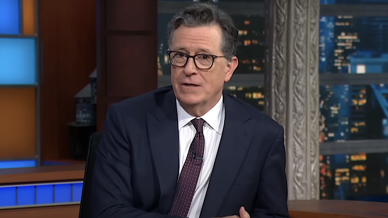 Stephen Colbert wearing a suit and tie while sitting at a desk hosting The Late Show with Stephen Colbert. 