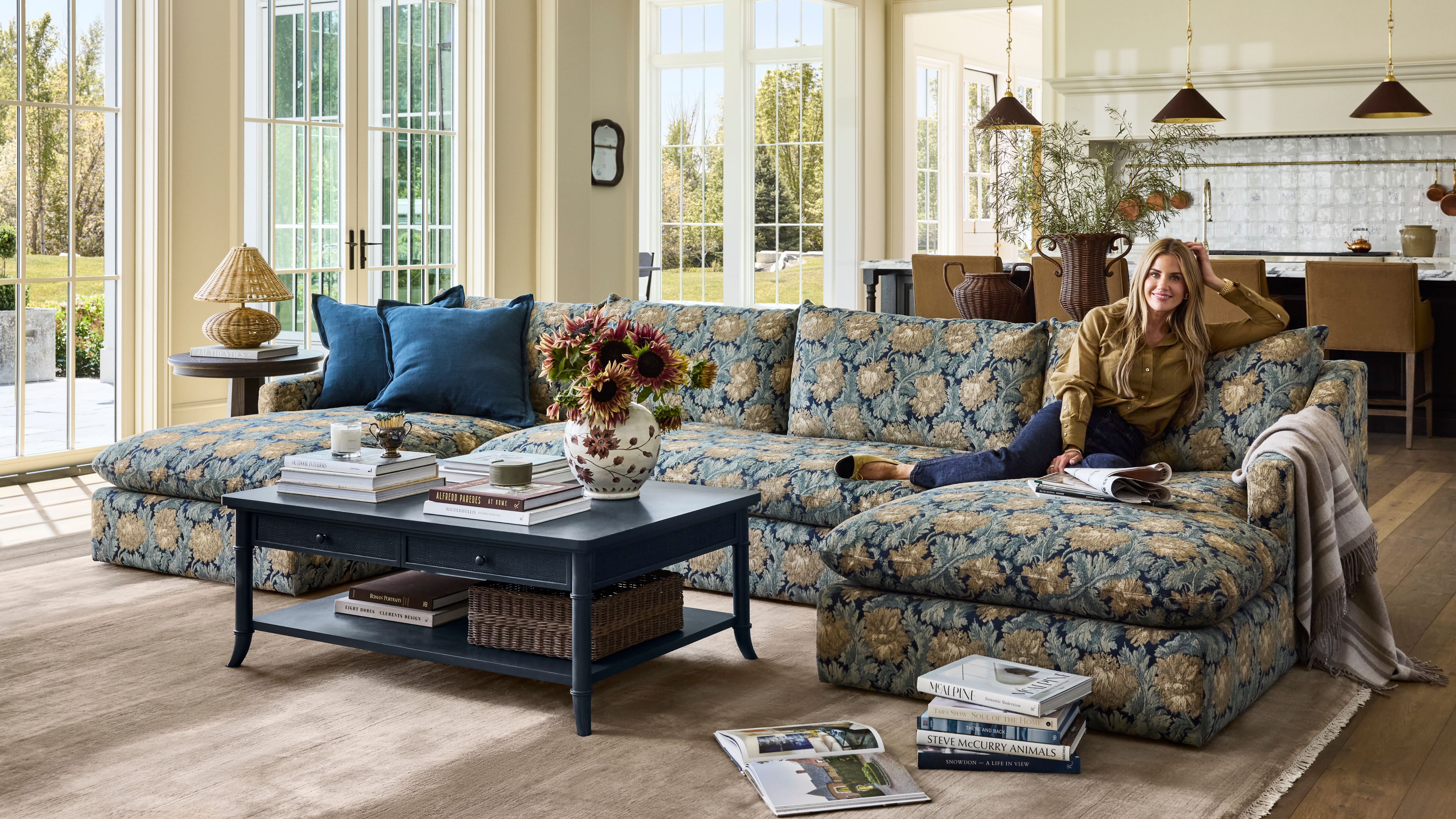 Shea McGee smiles on a floral jacquard sectional from her new McGee & Co. fall 2026 collection, which is styled with a navy storage coffee table, taupe rug, woven table lamp, and collected objects