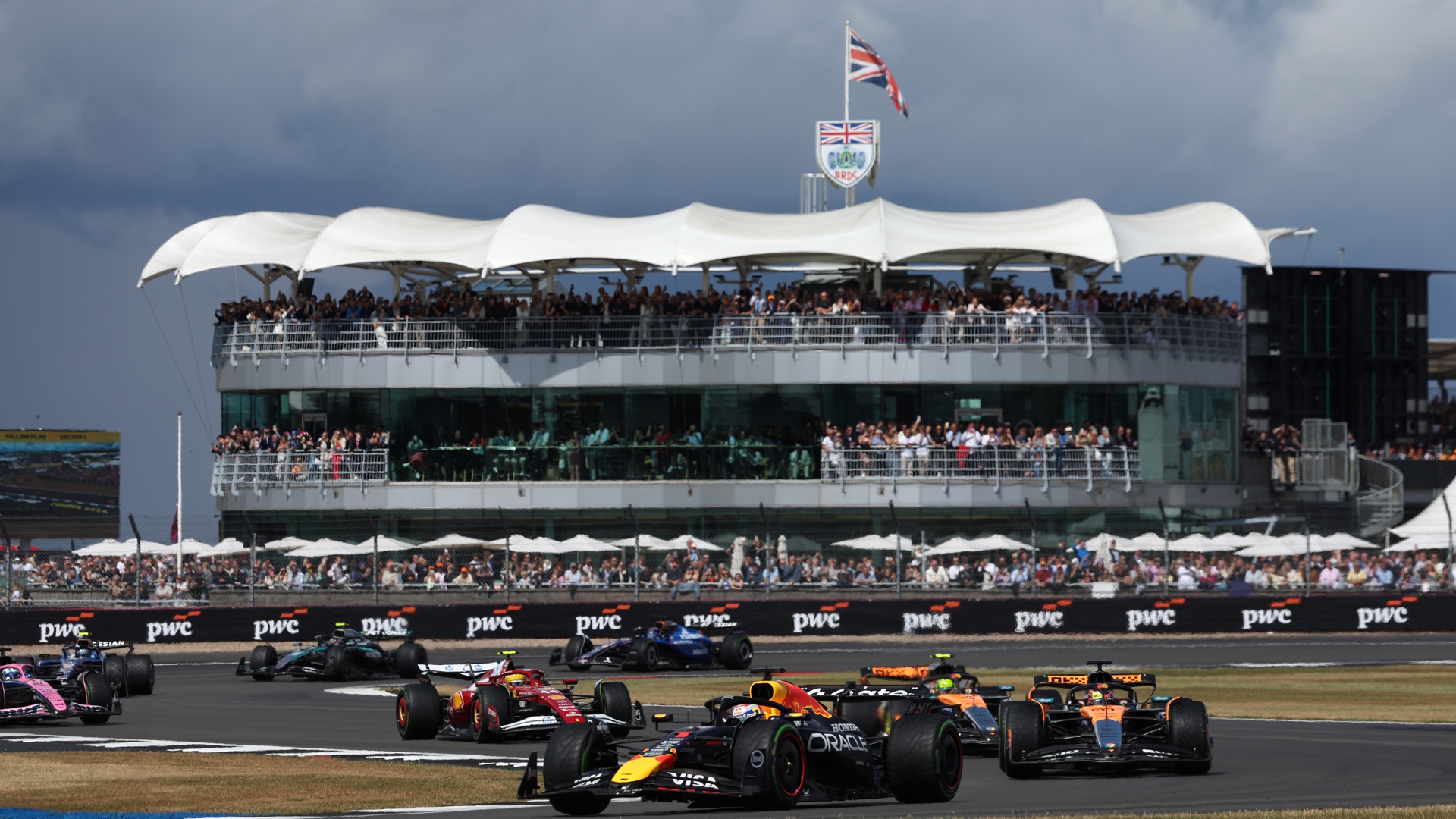 Formula 1 care on the track during the British Grand Prix at the Silverstone Circuit with a United Kingdom flag flying in the background.