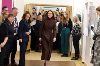 Kate Middleton wearing a long plaid coat walking through the hall of a hospital with staff surrounding her