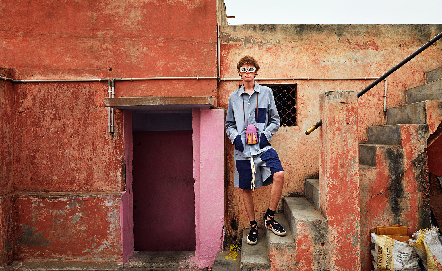 View of a male model wearing a light blue and dark blue shirt and shorts. He has sunglasses on, a bag around his neck and is standing on the steps outside a building with distressed red walls and a pink entrance