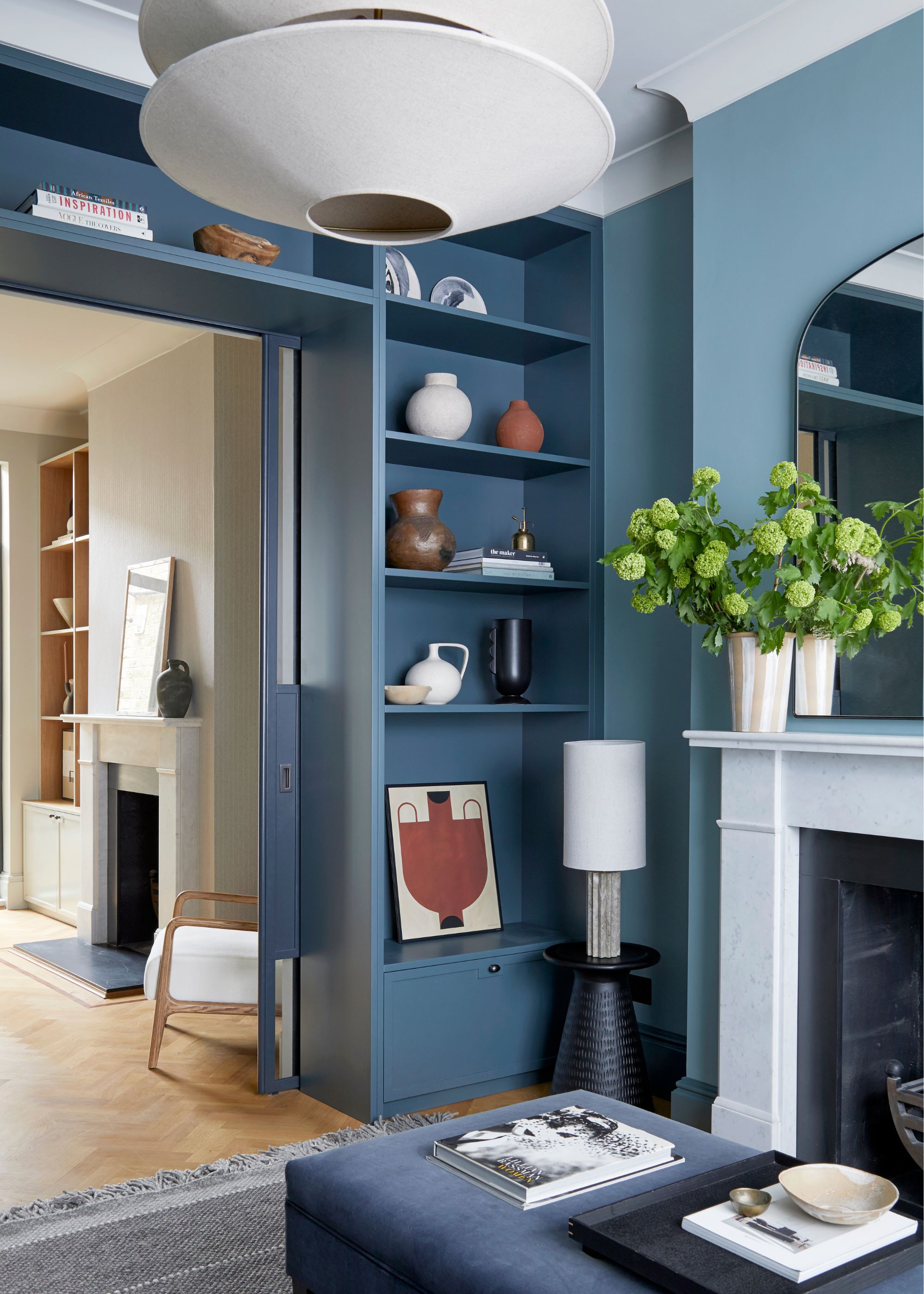 A traditional living room with warm blue walls, darker blue color-drenched shelving, a marble fireplace with a vase of green flowers, and modern decor.