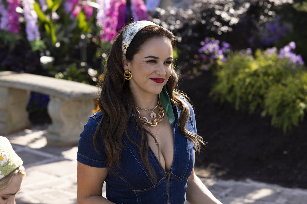 Still from Grosse Pointe Garden Society with Melissa Fumero as Birdie wearing a low cut denim jumpsuit with printed headscarf and gold chunky jewelry.