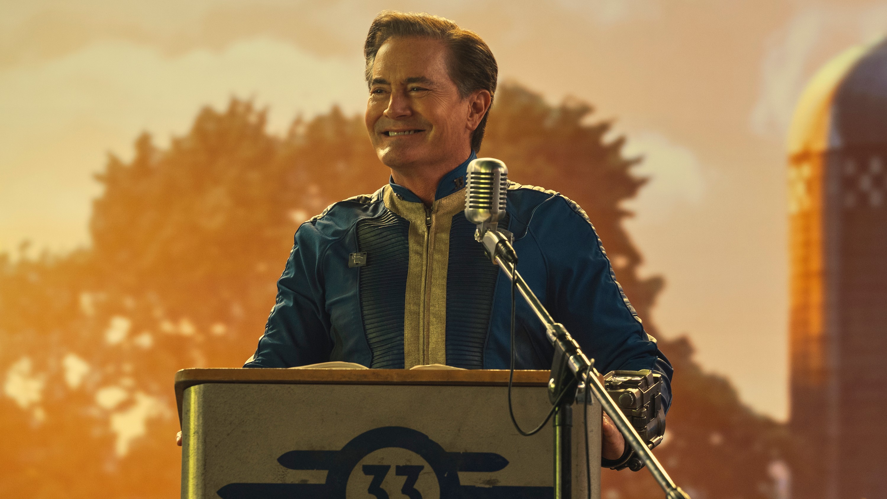 Overseer Hank MacLean (Kyle MacLachlan) in his Vault suit standing at a podium in Fallout.