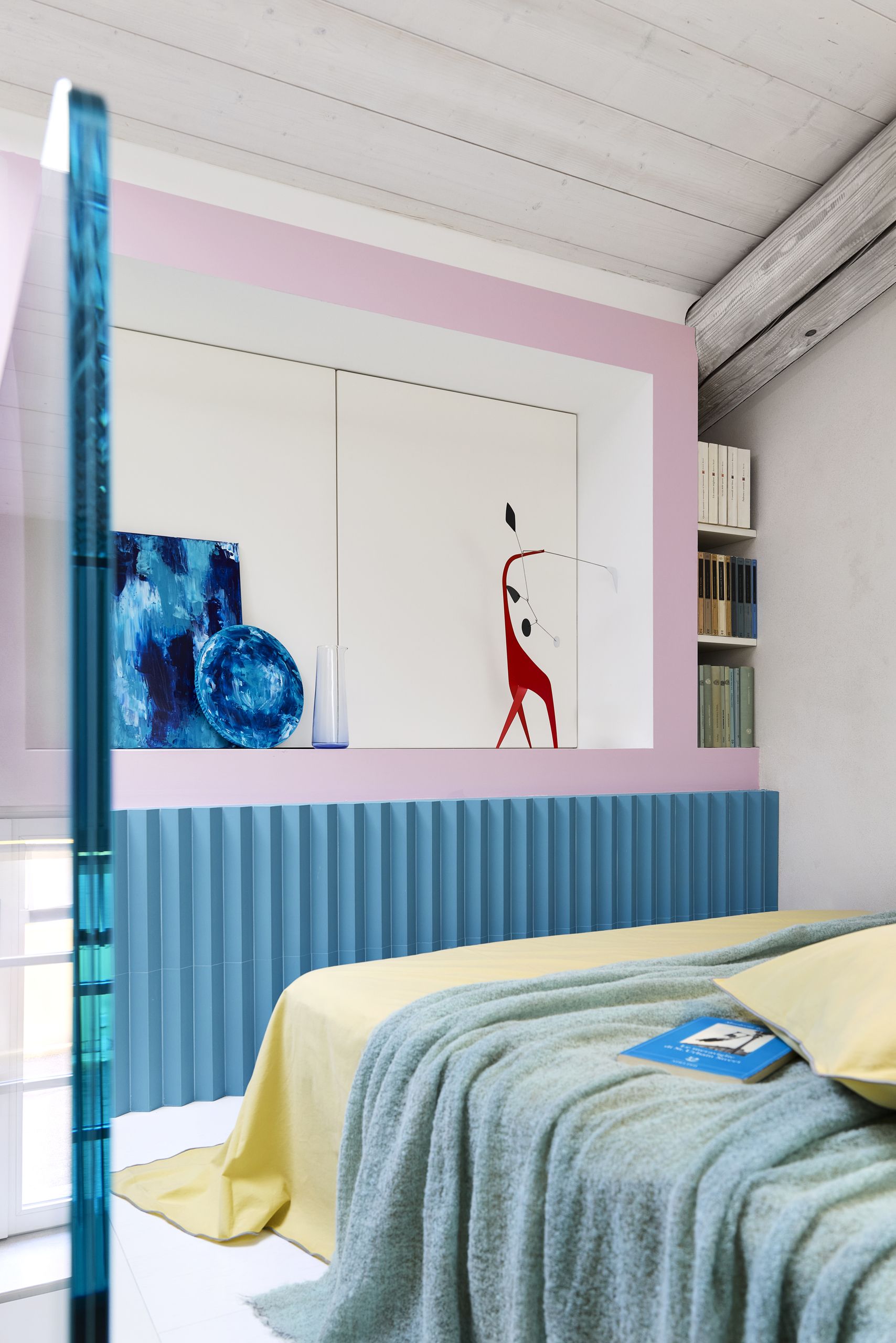 A loft bed with a pink and blue wall, a yellow bed, and blue leaning paintings
