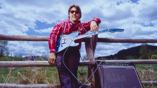 Steve Miller leaning on a fence holding a guitar, with an amplifier at his feet