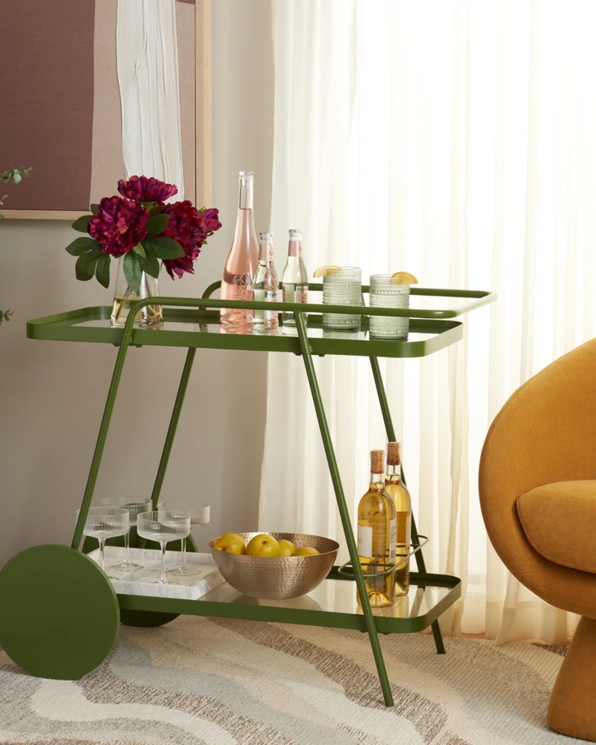 Image of a green metal bar cart with different drink accessories on it in front of a window with white curtains. There is a modern, ochre yellow accent chair next to the bar cart.