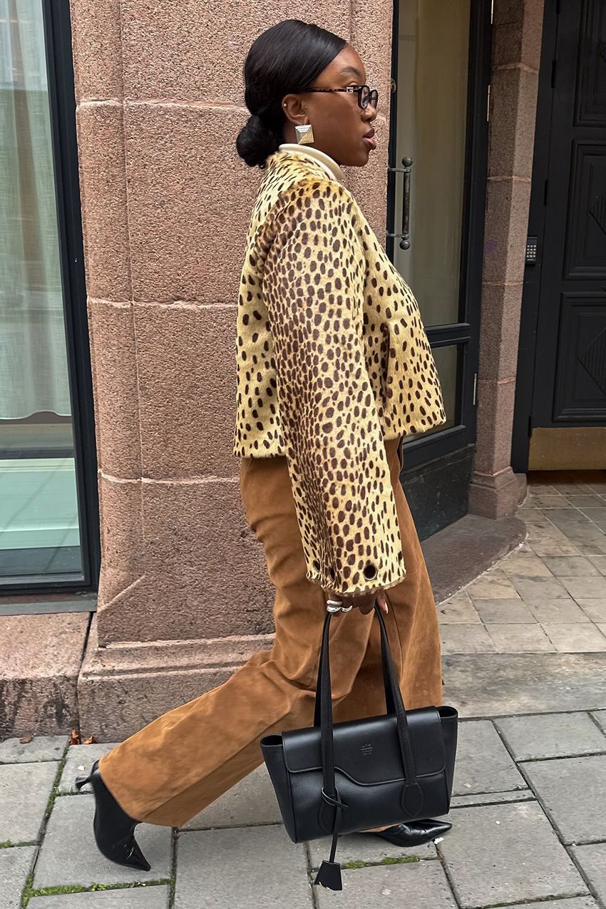 A woman walking outside down a sidewalk wearing a cheetah-print cropped calf-hair jacket styled with a white turtleneck sweater, light tan suede pants, black ankle boots, a black handbag, and silver jewelry.