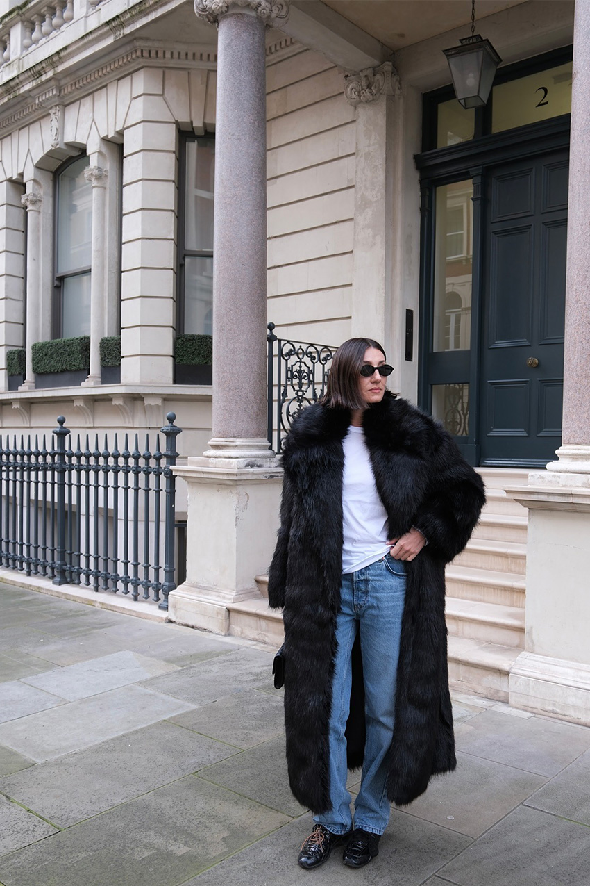 Denim and shoe combos shown in a street style photo of a woman standing on the sidewalk wearing a long black fur coat over a white t-shirt styled with slouchy straight-leg jeans, black patent leather racing sneakers, black sunglasses, and a black handbag