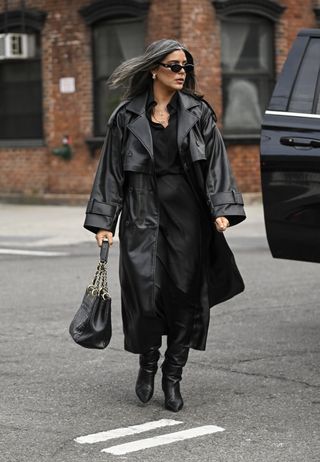 A woman is seen wearing a black faux leather trench coat, black gold chain bag and black boots with black sunglasses.