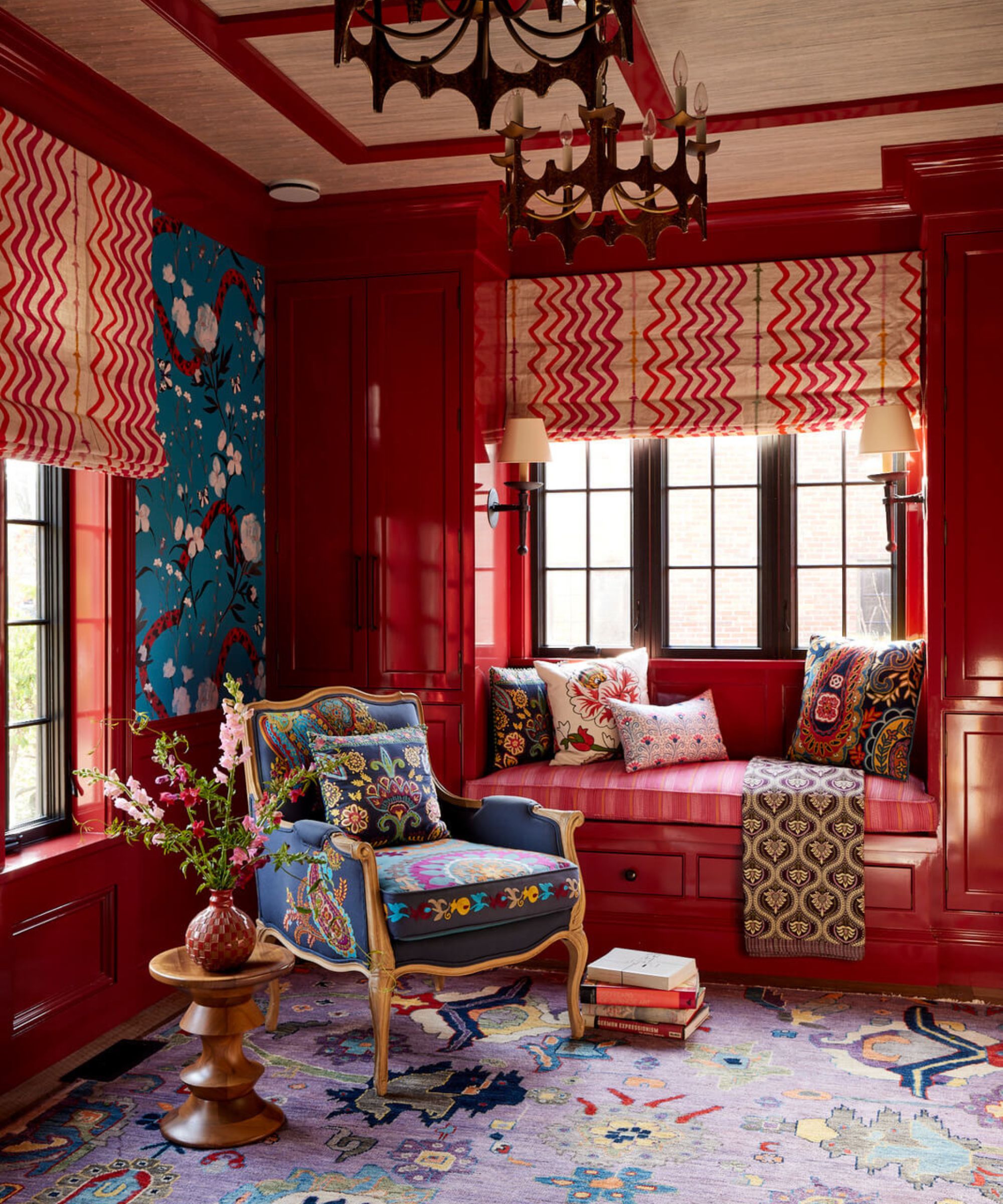 A maximalist room with glossy cherry-red lacquered walls and cabinetry, featuring a blue upholstered armchair with colorful embroidery, a pink window seat, and vibrant purple patterned rugs.