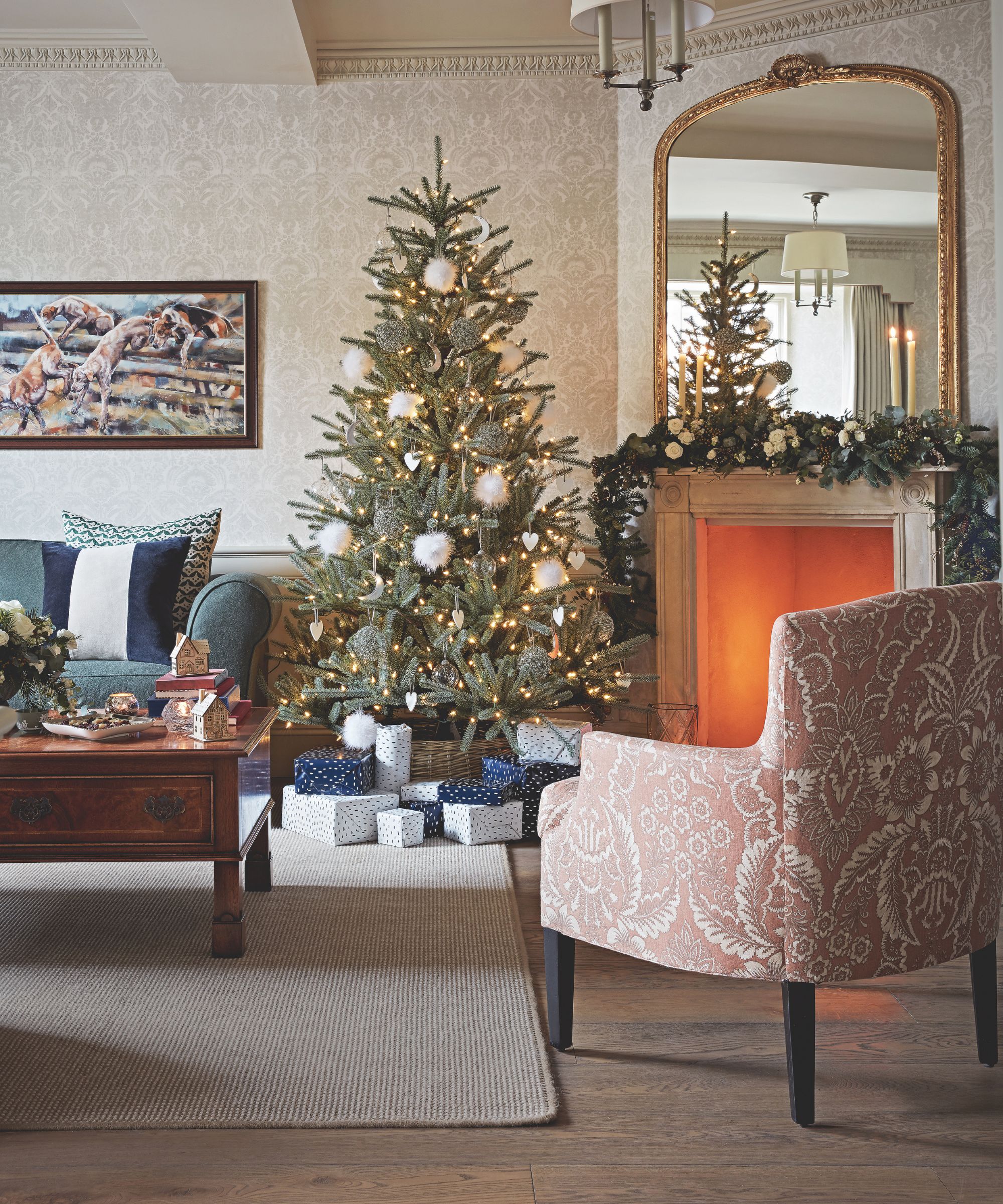 A cozy Christmas living room with a Christmas tree, lit fire in the fireplace, upholstered chair, rug, wooden coffee table and blue sofa.