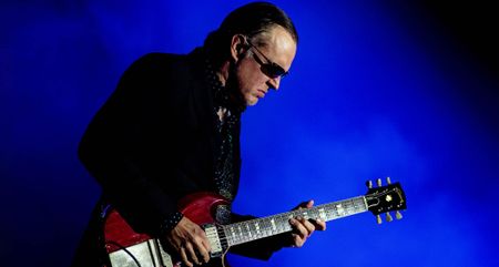 Joe Bonamassa wears a dark jacket, dark shades, as he plays his Gibson SG live against a dark blue background.