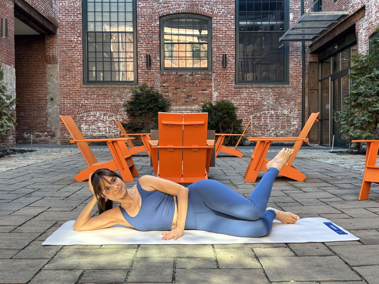 Pilates teacher Monica Borodin performs clamshell exercises. She wears a vest and leggings and exercises on a mat outside. Behind her we see a large brick building with long windows and a tablet and chairs in a courtyard. She is lying on her side with her legs stacked and knees bent at a right angle. Her knees are touching but the foot of her left leg is elevated and pointing towards the sky.
