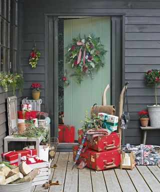 a porch with dark grey walls and green door, decorated with country christmas decorations including wreath on door, red vintage suitcases, wrapped presents and red and green plants
