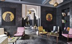 A hotel sitting area with different coloured wood cloth covered chairs, wooden coffee tables, dark grey walls, wall paintings, a book shelf, patterned floor tiles and a long corridor. 