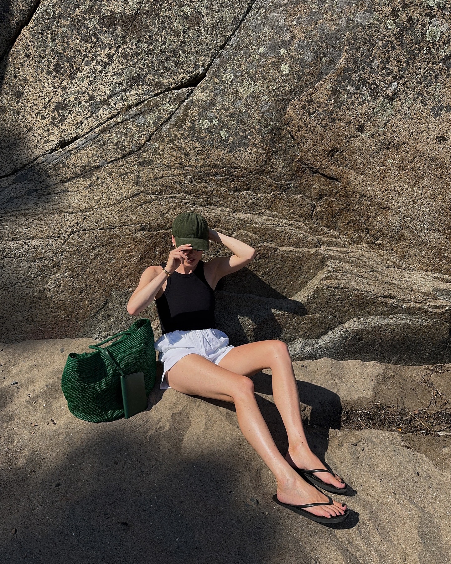 Woman wears black tank top, white shorts, black flip-flops, green woven bag
