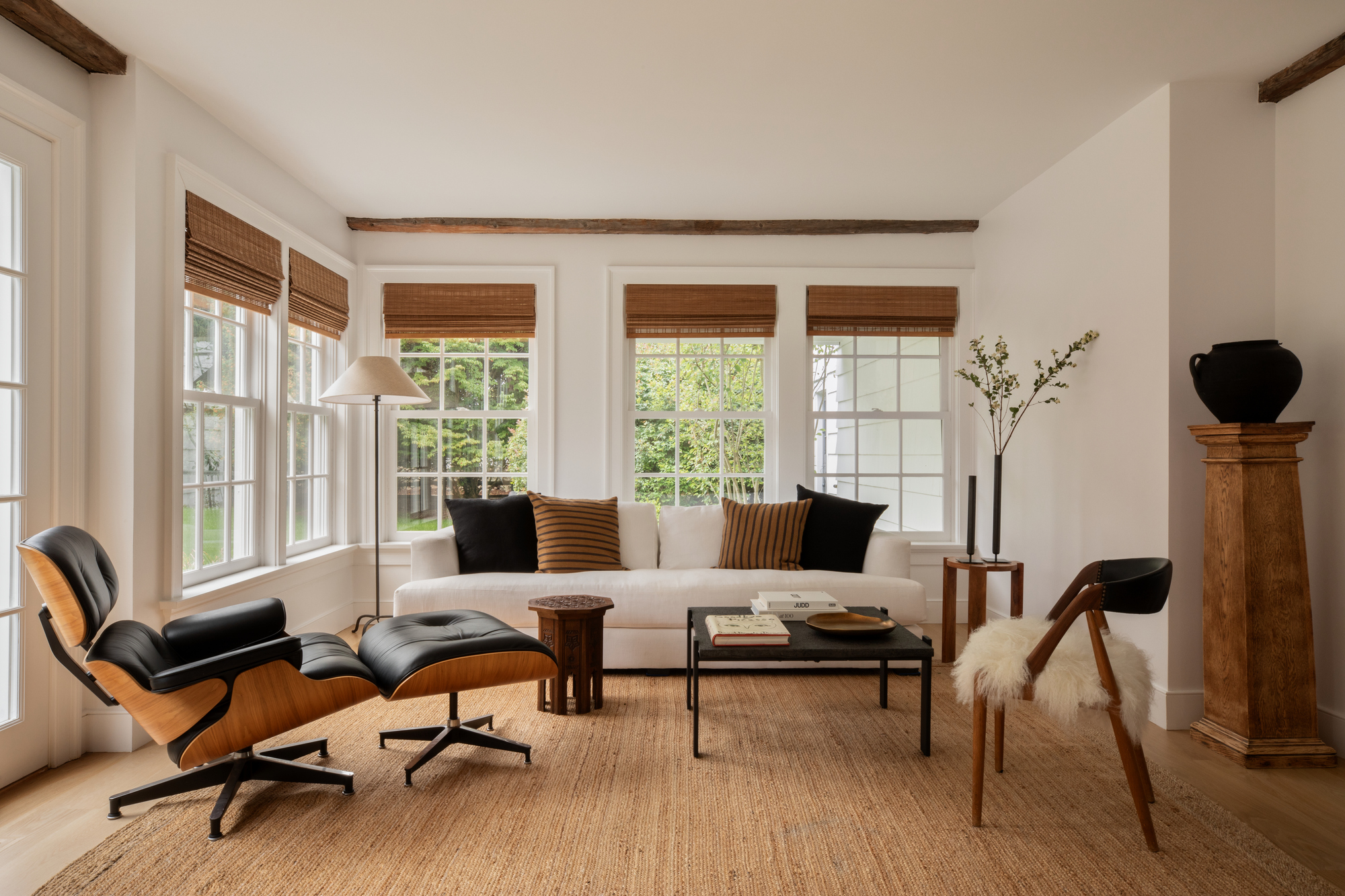 modern living room with bamboo blinds, a white sofa with caramel and black throw pillows, an eames lounge chair, a square coffee table and side table, an armchair with a sheepskin, a pedestal with a vase styled on top, and a jute rug on the floor