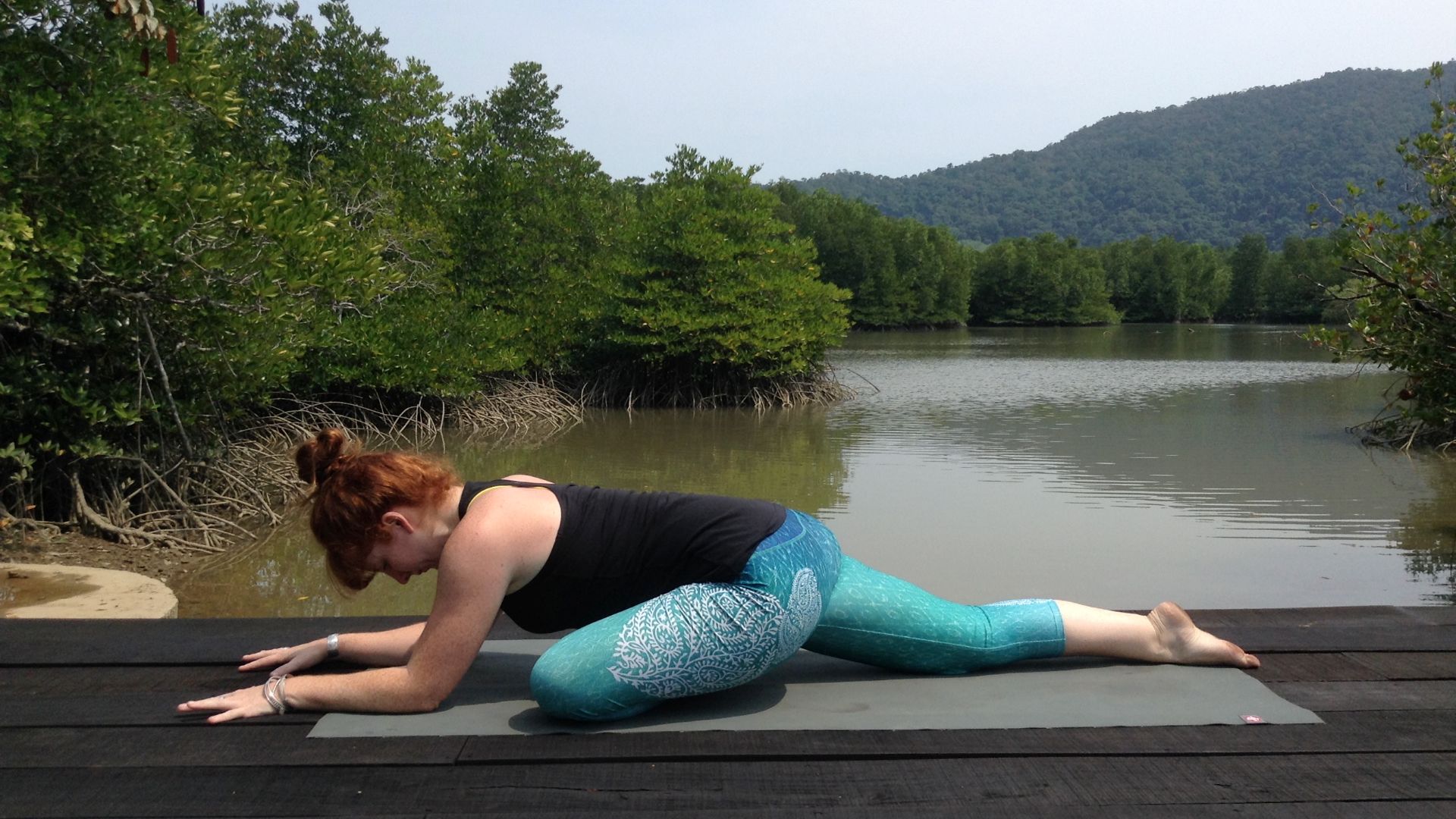 Yoga teacher Alison Reaume in pigeon pose. She is on a wooden platform with a lake and trees in the background
