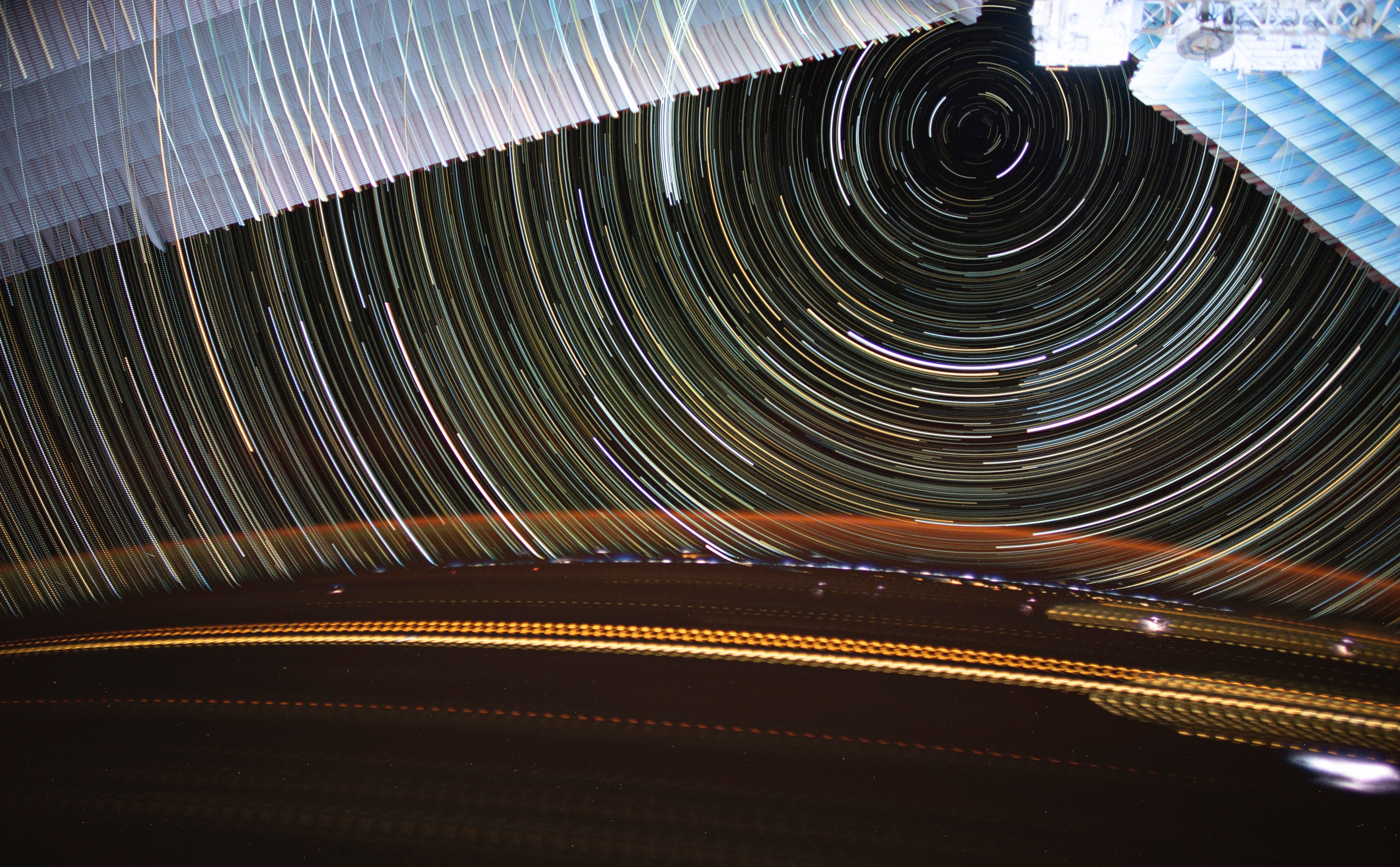 Estelas de luz cruzan la Tierra mientras las estelas de estrellas giran sobre nosotros en esta impresionante fotografía de larga exposición tomada por la astronauta de la NASA Christina Koch en la Estación Espacial Internacional.  En la parte superior del marco se encuentran los paneles solares de la estación espacial.  Abajo, los relámpagos se esparcen por la superficie de la Tierra, mientras que un resplandor naranja ilumina la atmósfera.