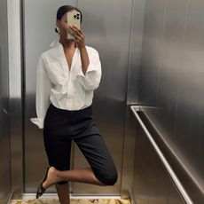 French woman wears black capri pants, white button down and black sandals in a mirror selfie. 