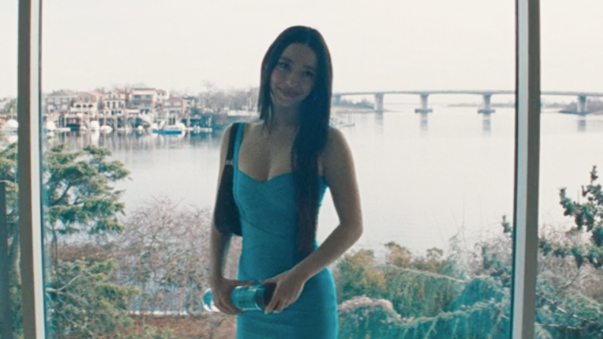 Mikey Madison smiling in a blue dress standing in front of a big window overlooking water.