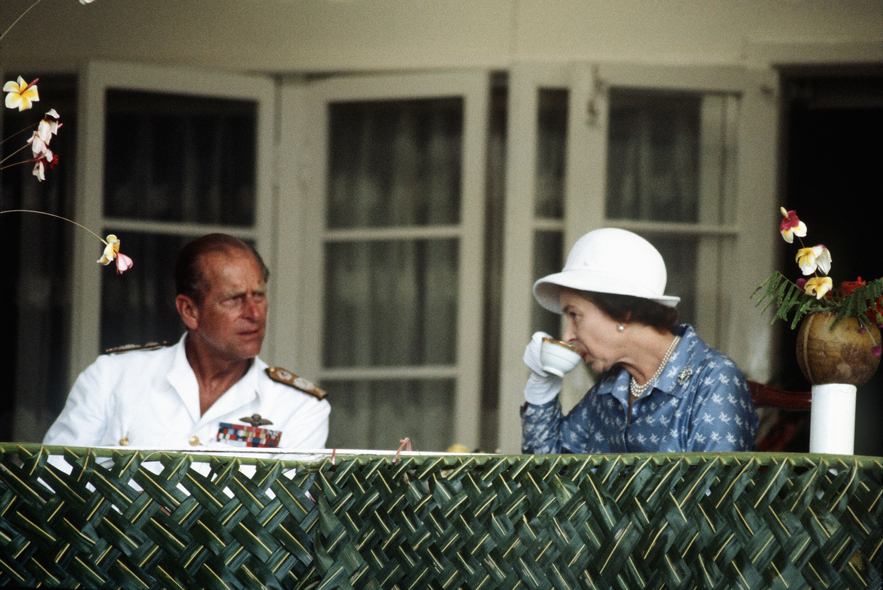 Queen Elizabeth drinking tea in a blue coat with Prince Philip, dressed in a white naval uniform