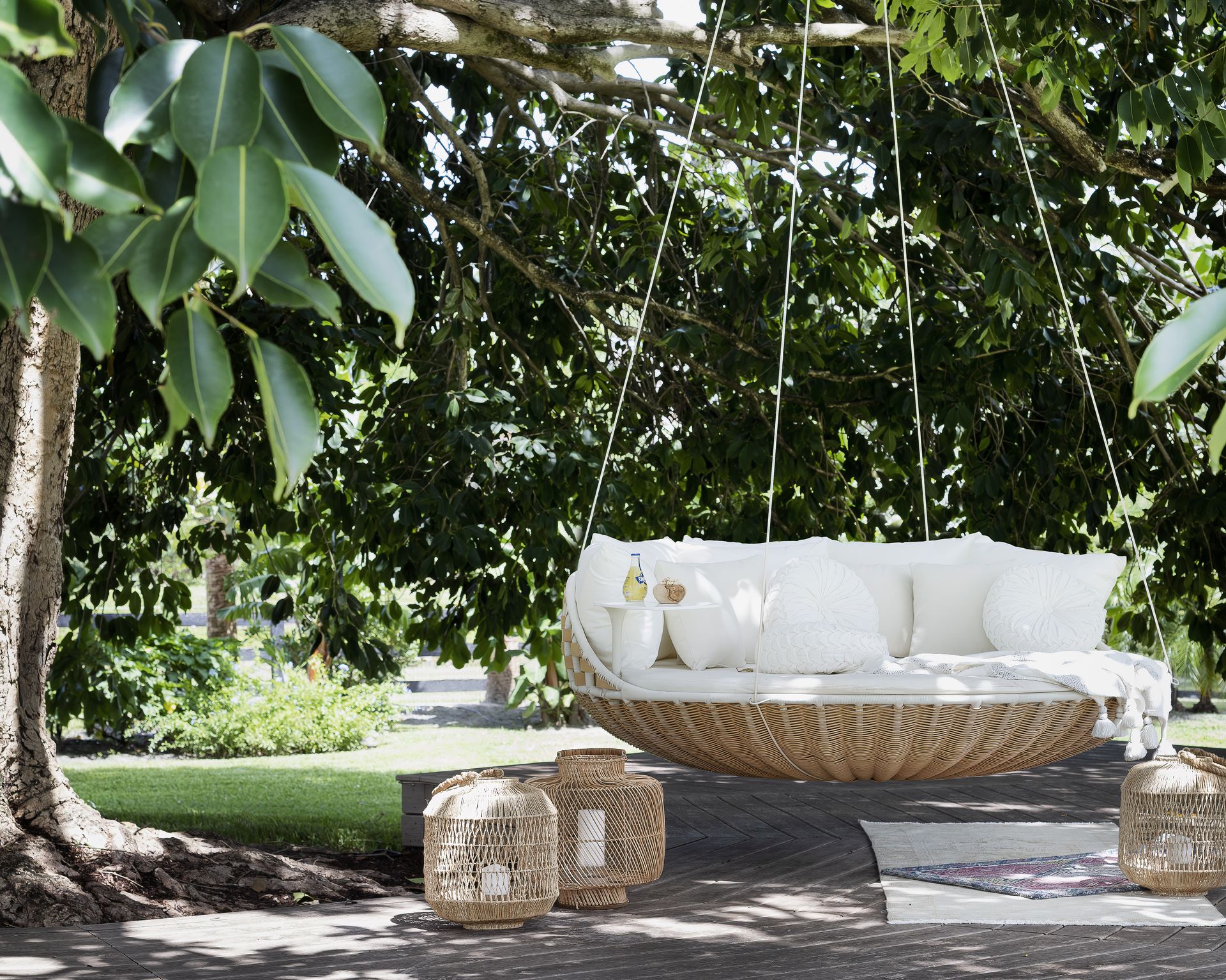 A lush garden, with a hanging round swing under a large tree with vast foliage overhanging, and floor lanterns placed either side of the swing