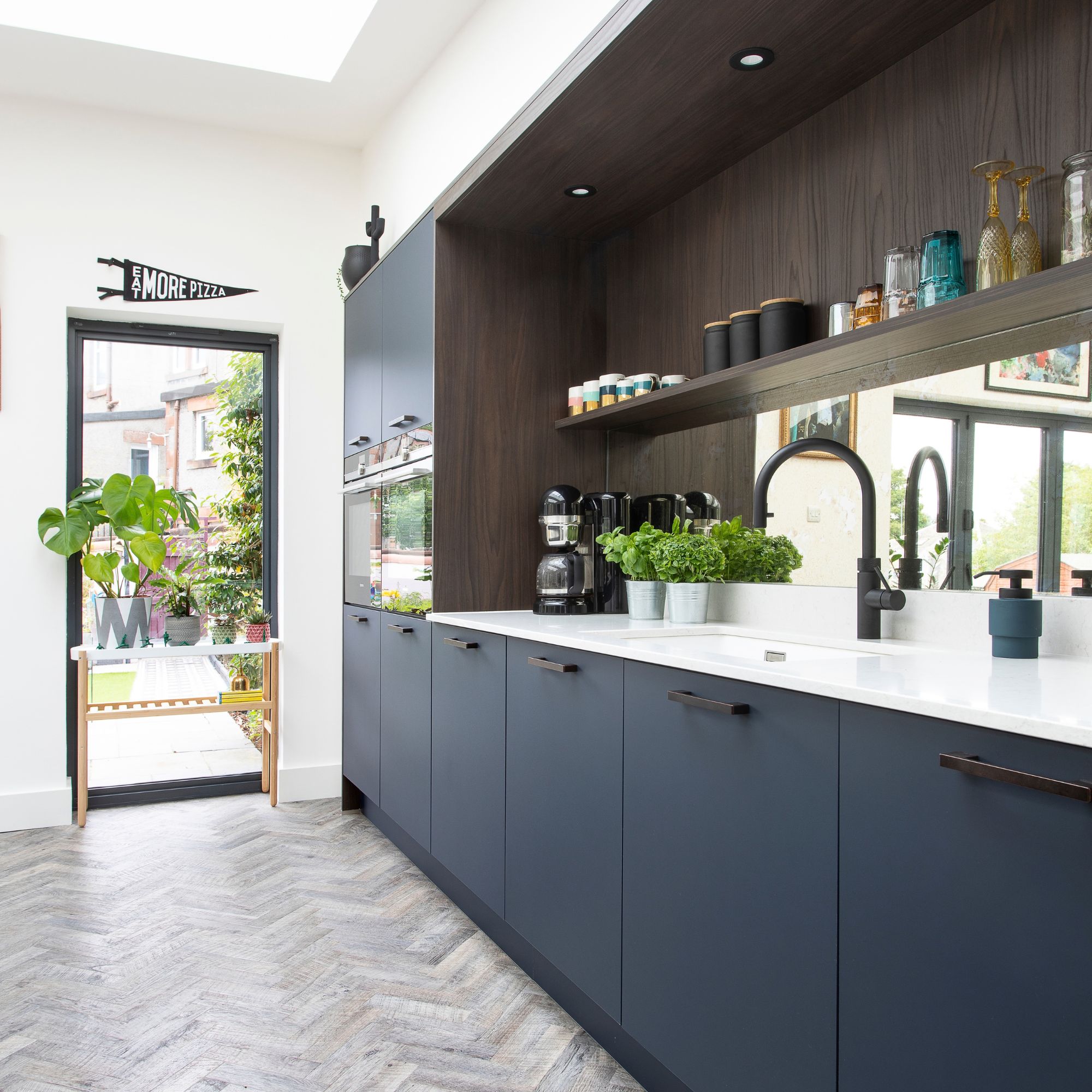 Kitchen with blue cabinets and grey flooring, with a dark wooden shelf above white worktops