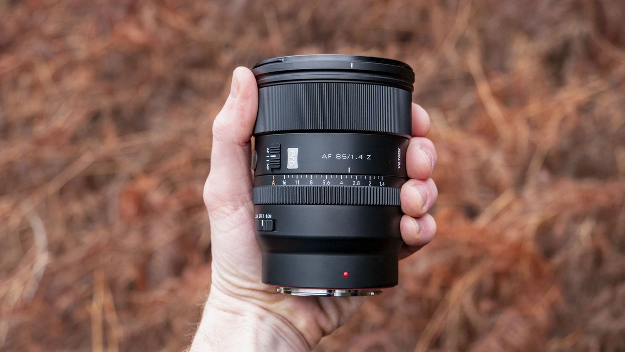 Viltrox AF 85mm F1.4 Pro lens in photographer's hand, outdoors and with bronze-colored ferns in the background