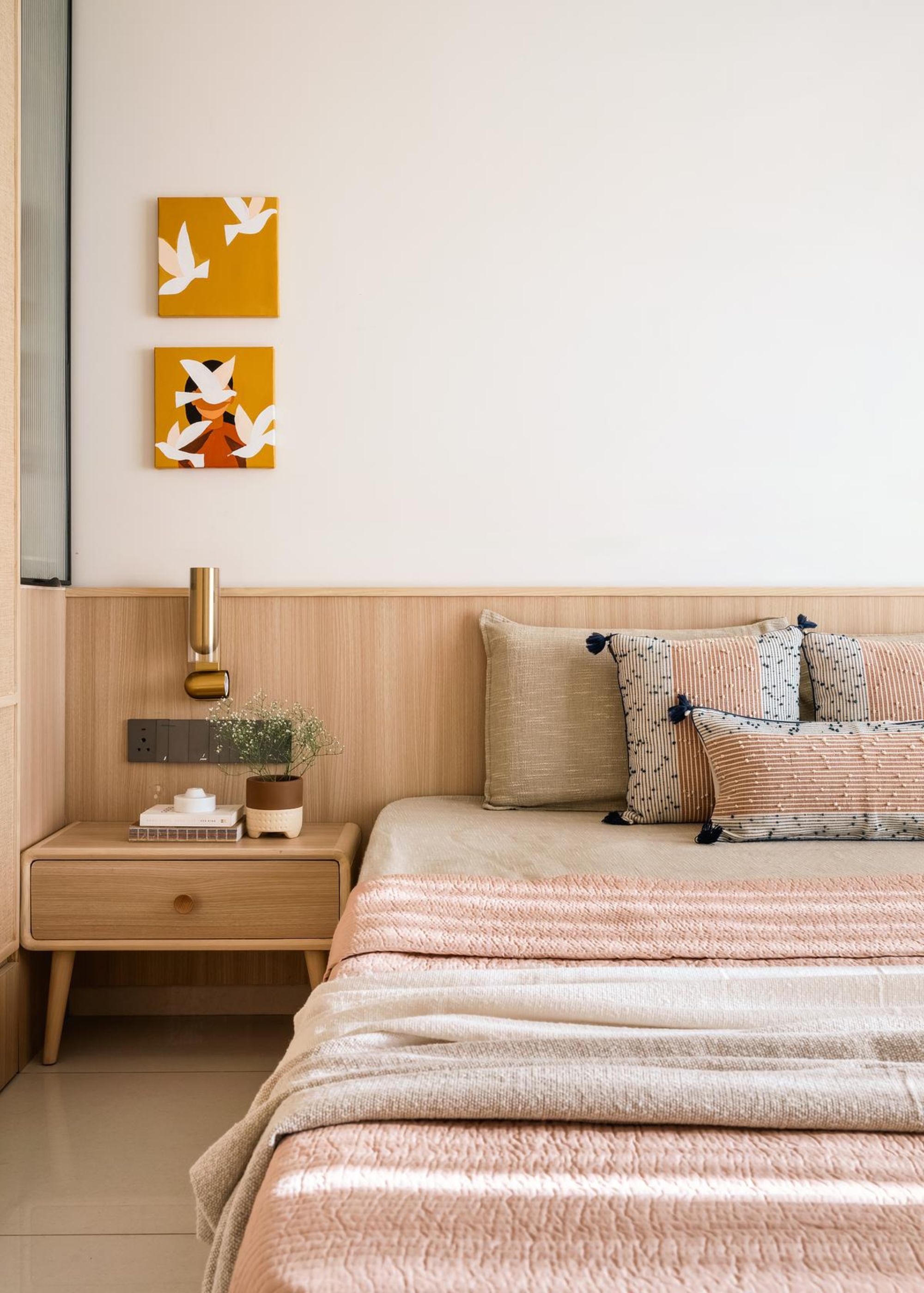 A view of the corner of a double bed with pillows and blankets on top. Next to the bed is a side table and above the table is a wall light as well as art hanging on the wall.