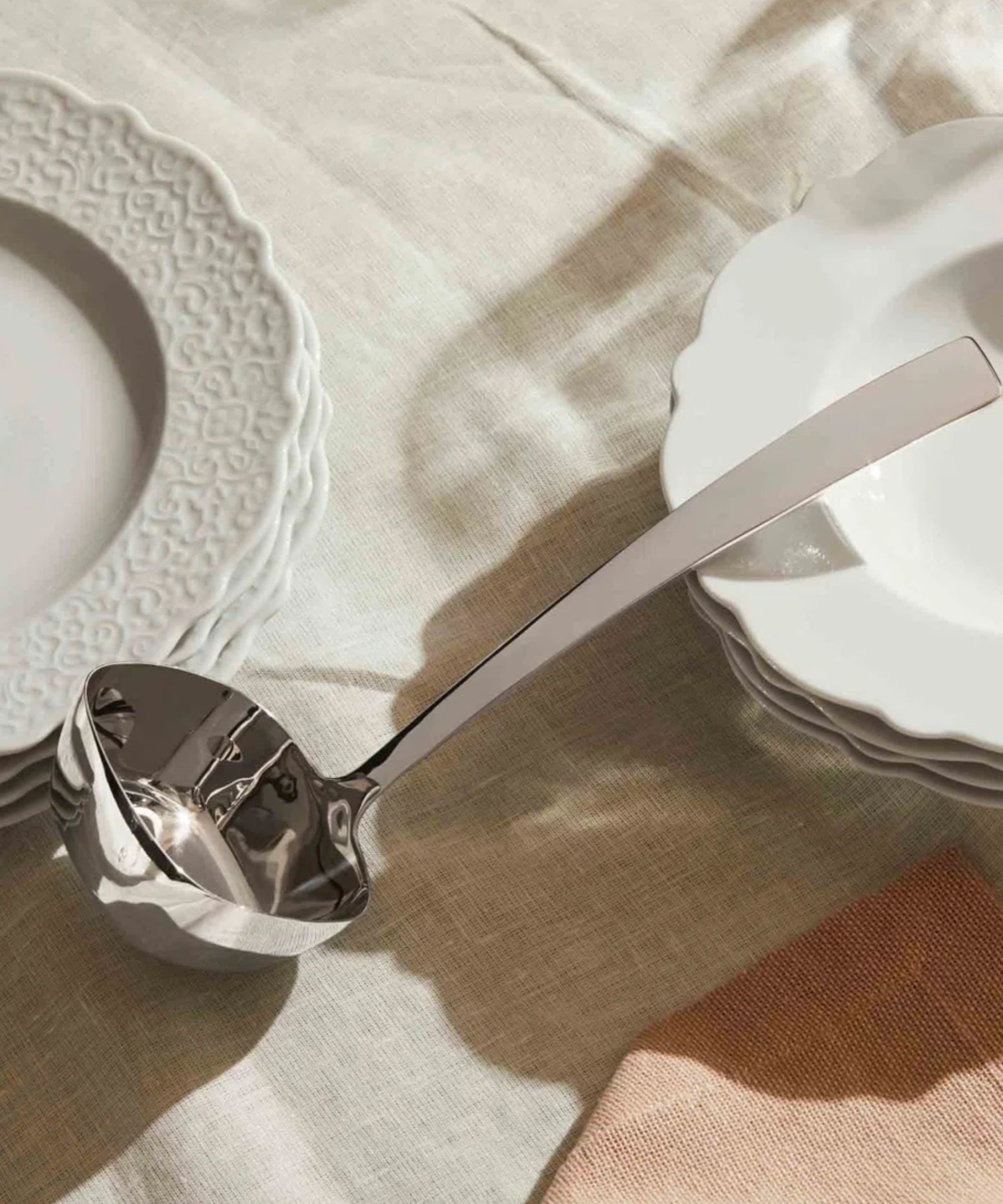 Silver square ladle on top of white tablecloth with stacked white scalloped plates