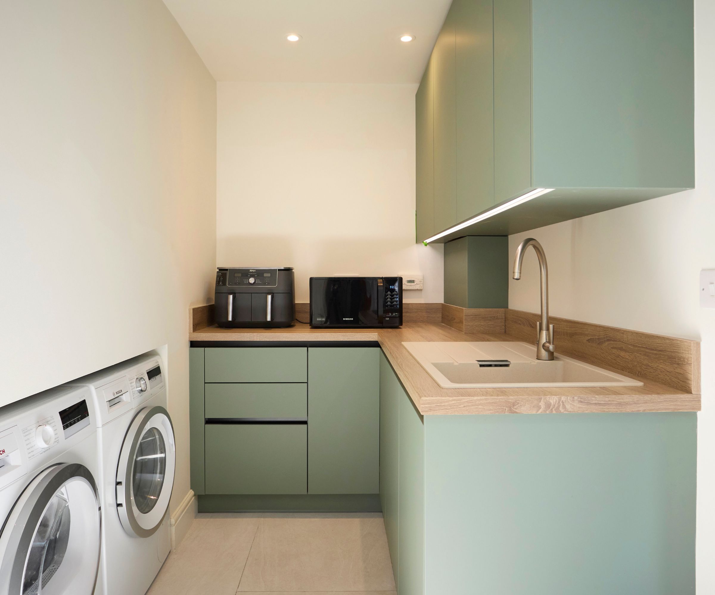 A compact utility room with sage green handleless cabinetry, oak worktops, a white composite sink, washing machine and dryer, and large format cream floor tiles.
