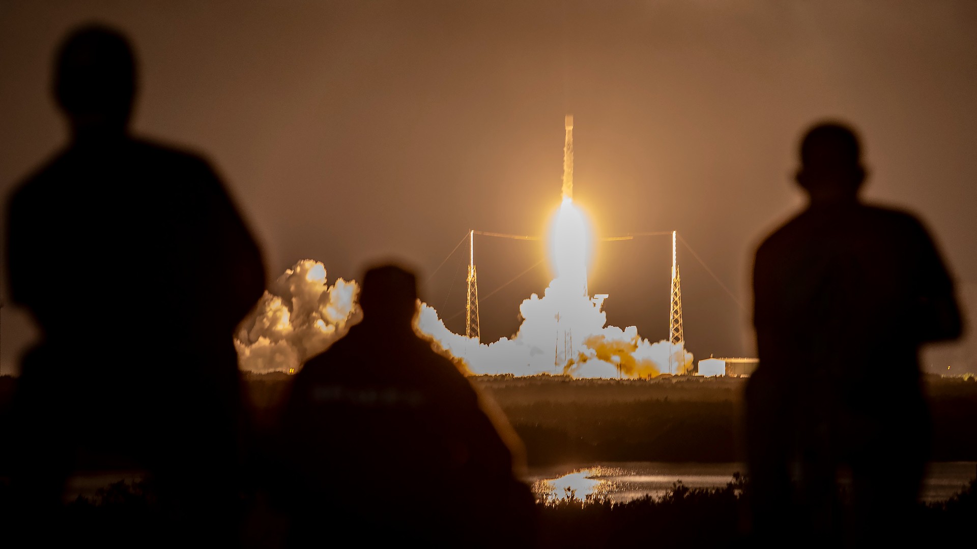 A rocket takes off into the night, lighting up the sky.  The faded silhouettes of people appear in the foreground.