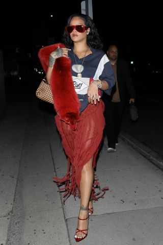 A photo of Rihanna pairing a sheer red skirt and navy polo with strappy red sandals and a ruby fur stole.