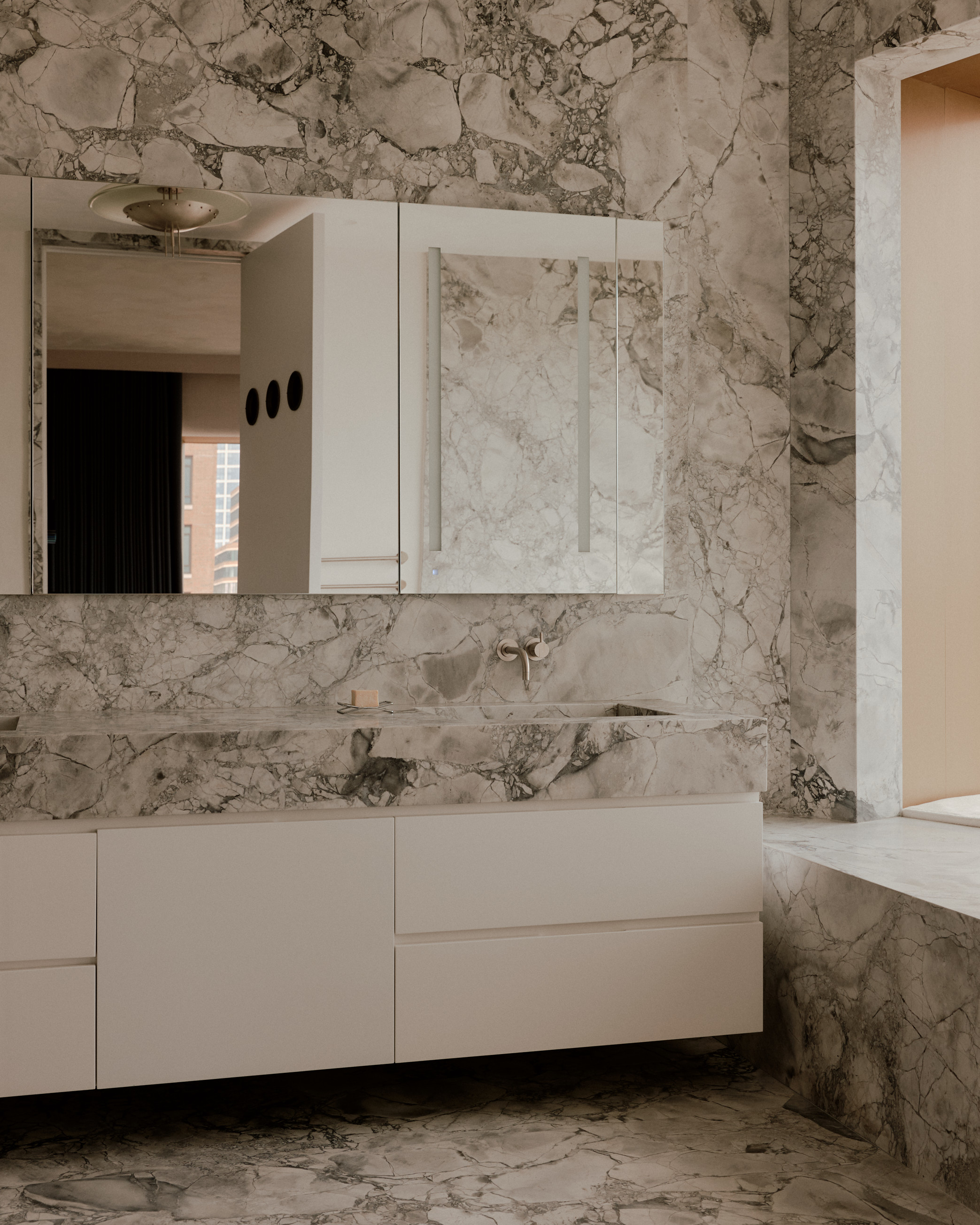modern bathroom drenched in marble walls, floor, built-in bath and vanity with white cabinets and cabinets behind mirror