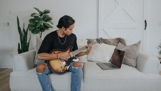 Man sits on a sofa with his guitar viewing a lesson on his laptop
