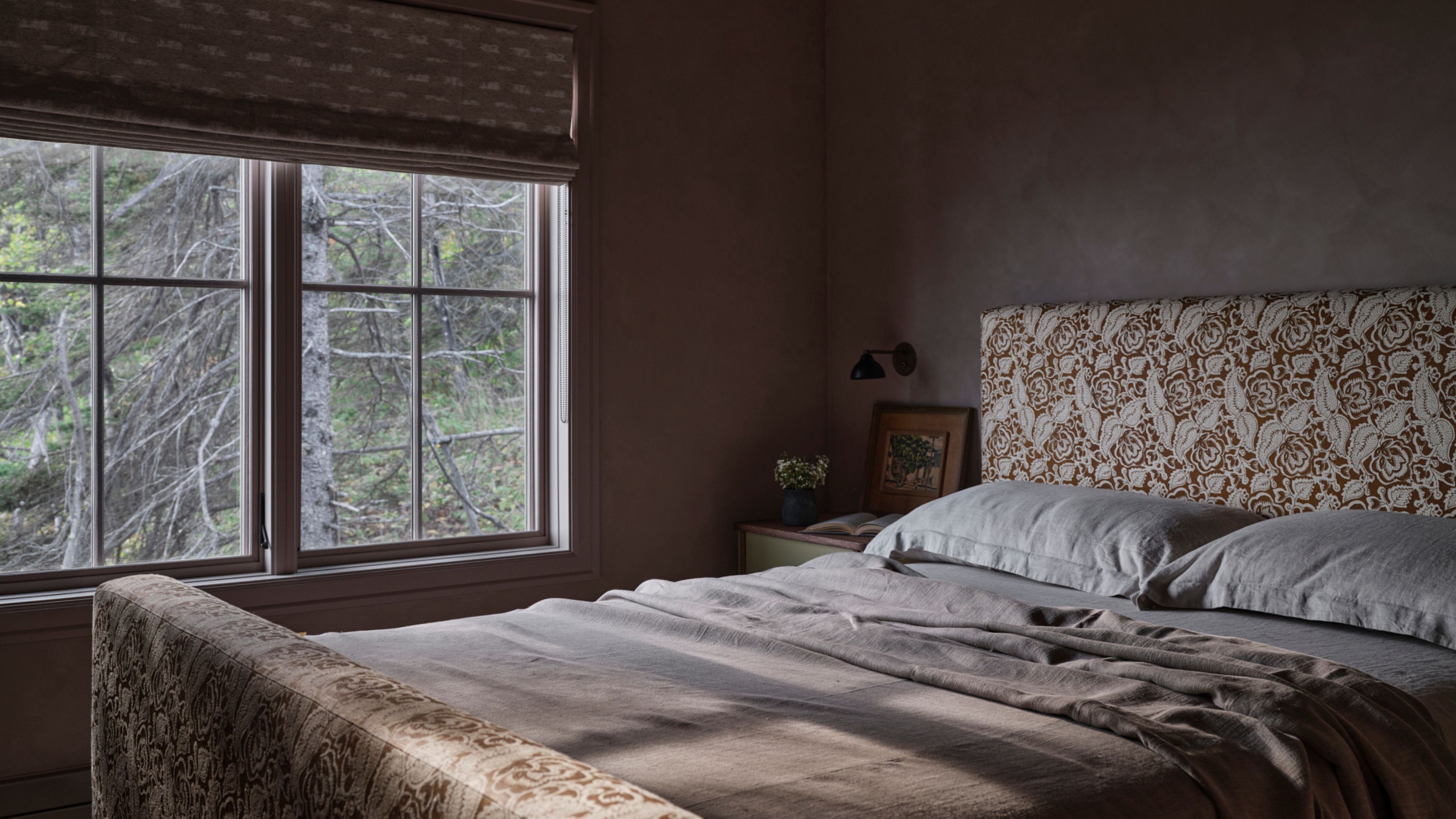 a moody plastered pink mauve bedroom with a floral upholstered headboard and window with a forest view