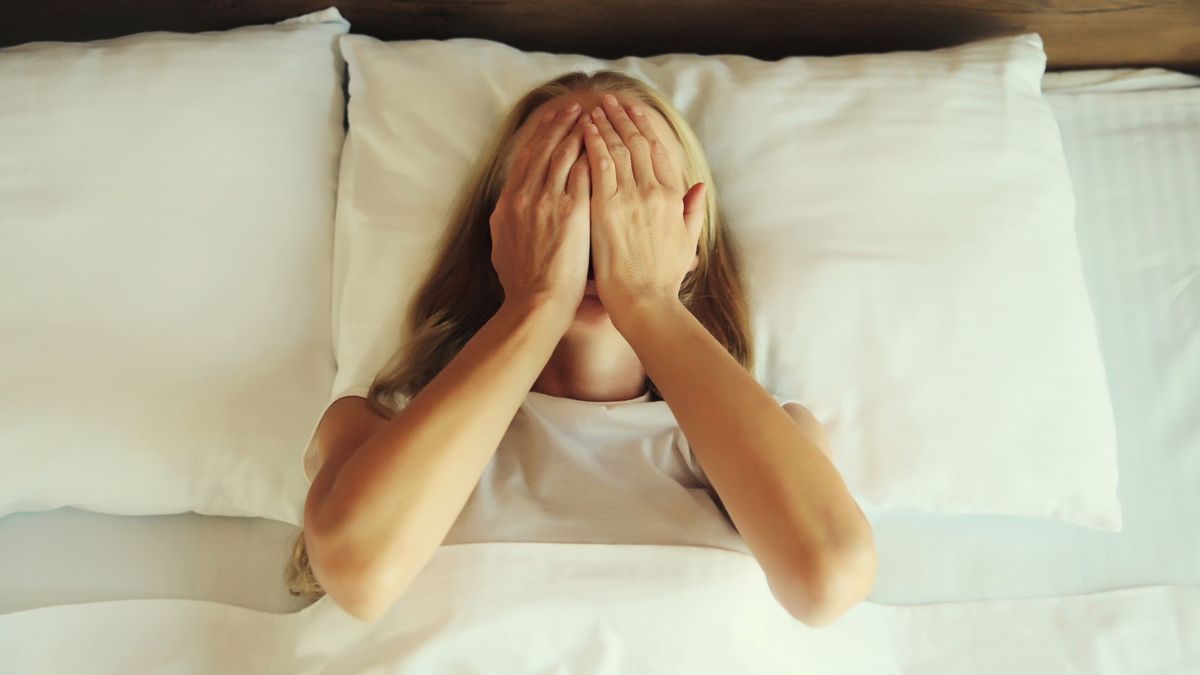 The image shows a blond-haired woman laying in bed and covering her face with her hands