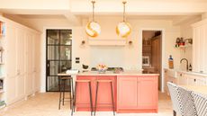 Kitchen island trends are so chic. Here is a peachy pink kitchen island with a quartz surface with a fruit bowl on top, two white globe pendant lights above it, and three wooden stools tucked under it