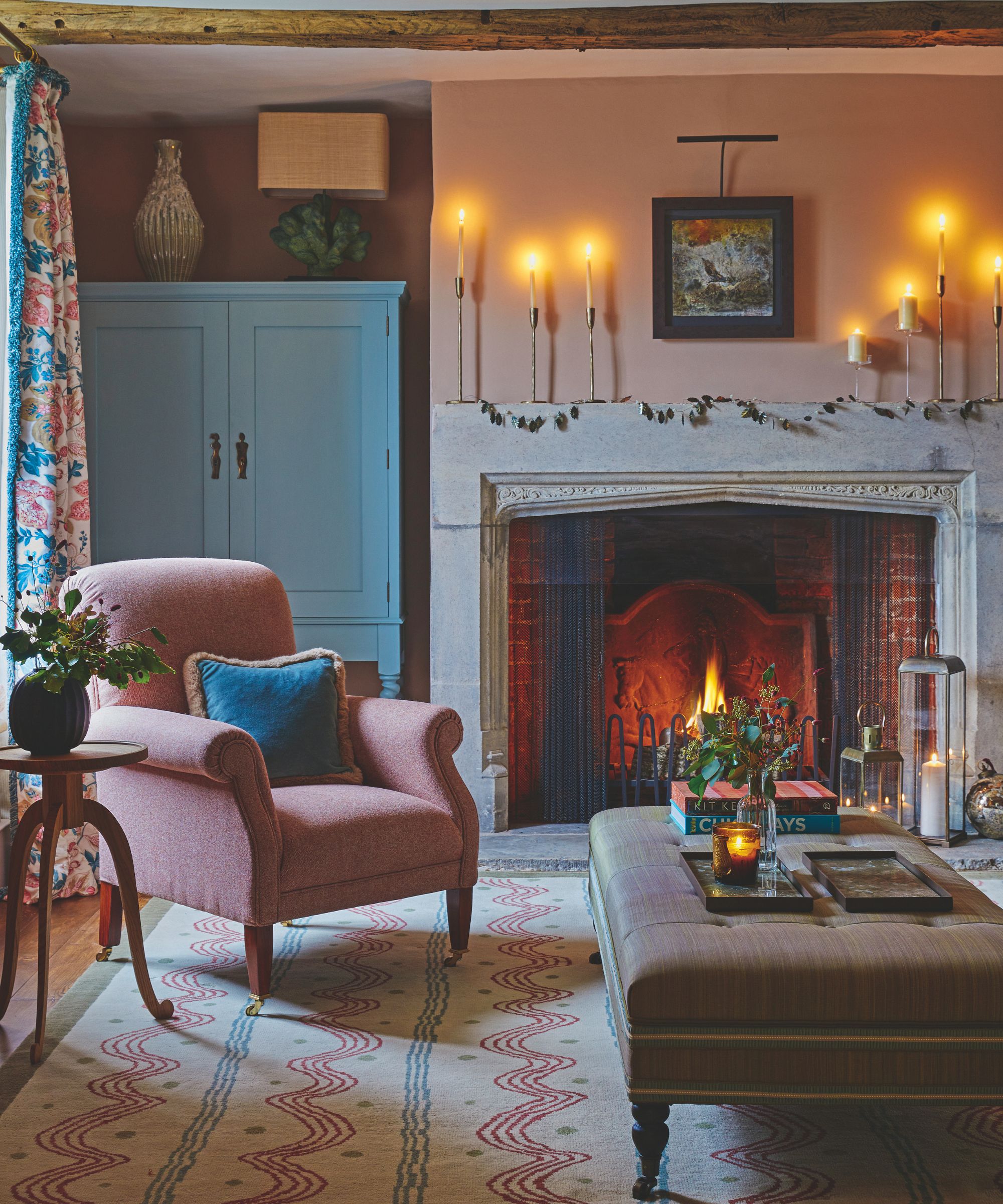 A cozy living room with a lit fireplace, candles, pink walls, a patterned rug, upholstered ottoman and pink chair.