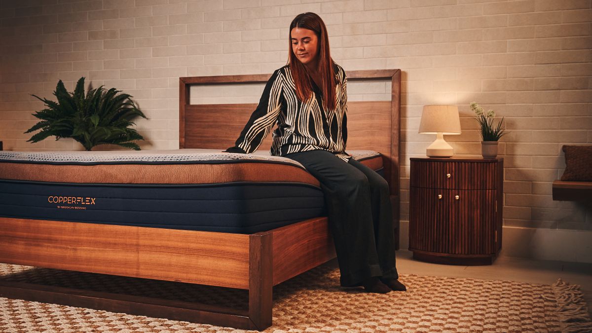 A woman in black and white silk pyjamas sitting on edge of Brooklyn Bedding Memory Foam Mattress on wooden bedframe in warmly lit sleep studio