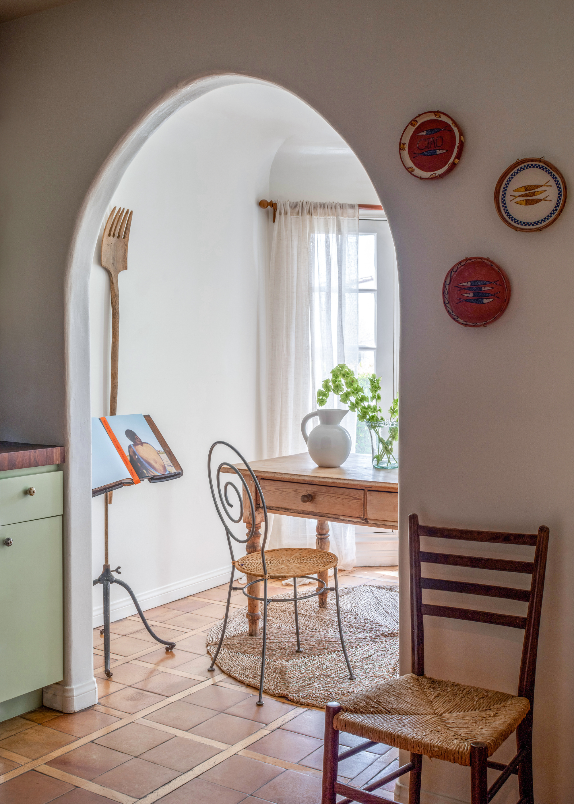 An arched doorway leading to a bright room with a wood table, a woven rug, a metal rattan stool, and a vintage book holder