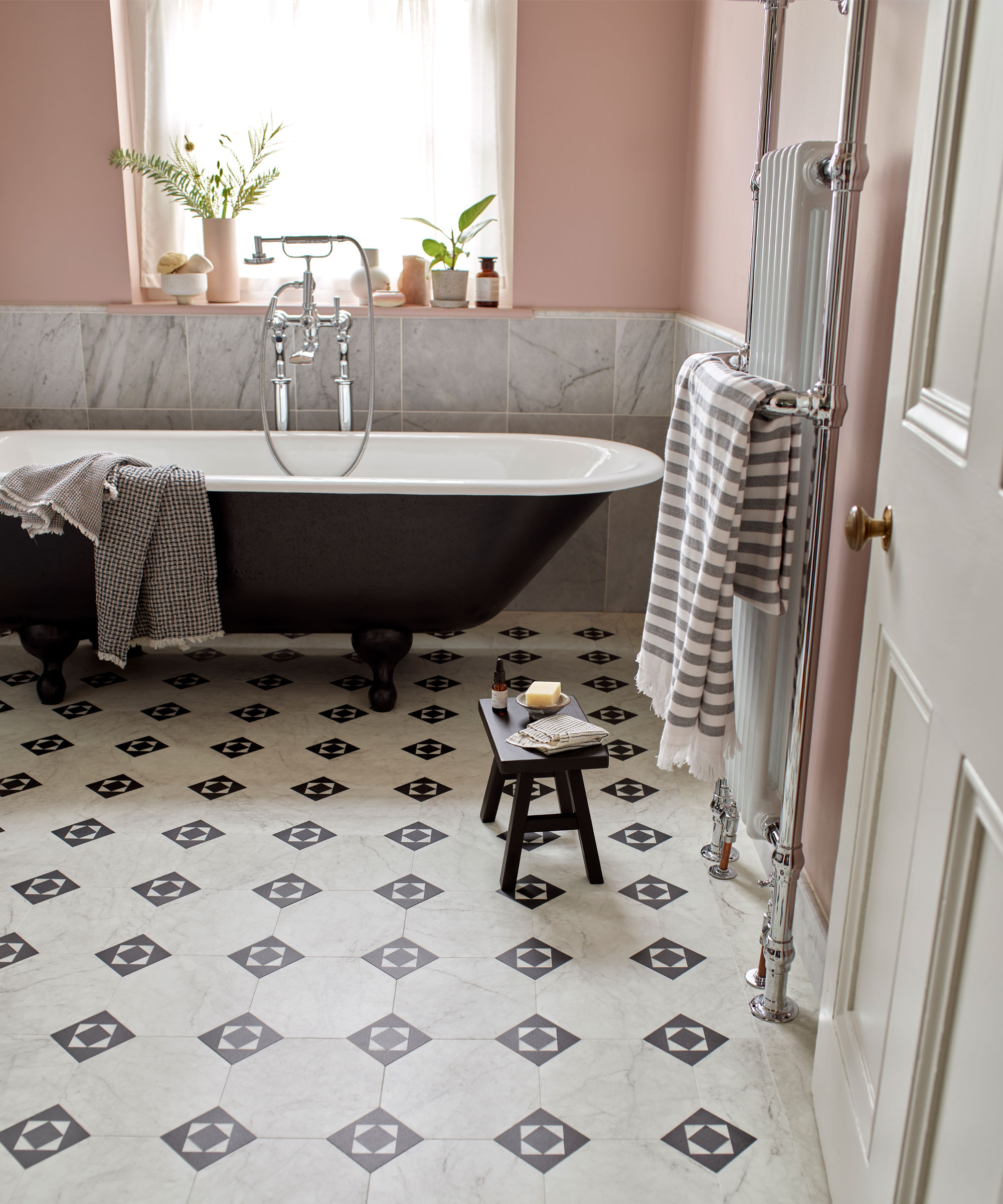 Bathroom with laminate tiles in traditional Victorian pattern in marble and black, marble wall tiles mounted halfway on wall, rest of wall painted a light pink, black bathtub, wooden stool, towel rail, window shelf with decorative vases, plants and toiletries